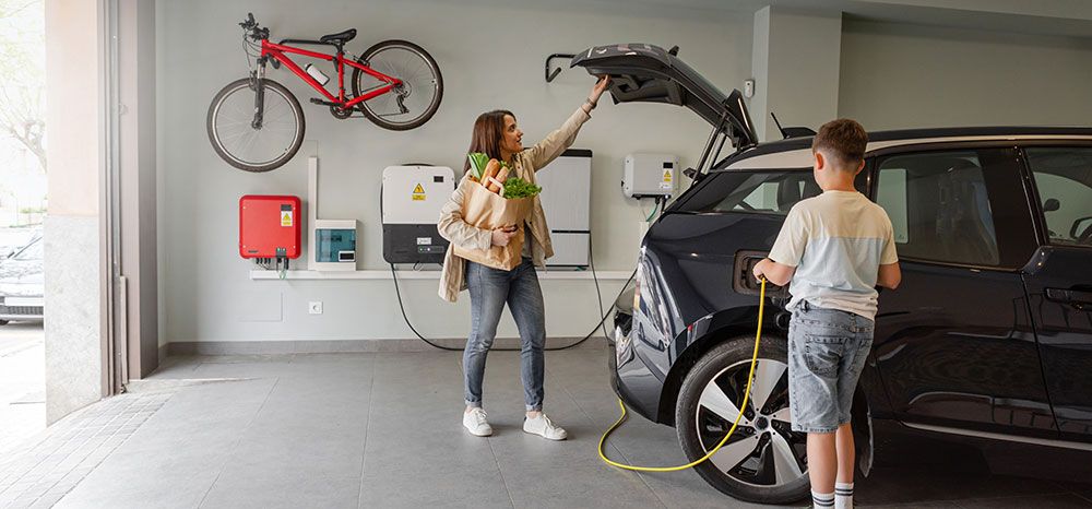 Kvinna och barn vid elbil som laddas i garage med laddbox och solpanelsystem.